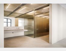 Moderner Sanitärbereich mit Betonwaschrinne und mehreren Armaturen vor einer metallverkleideten Trennwand. Rechts sind Kabinentüren in bronzefarbenen Metallpaneelen, darüber eine Rasterdecke mit linearen Leuchten und links ein Fenster.