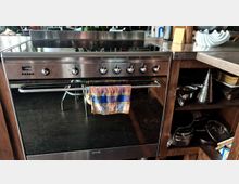 Moderne Küchenszene mit einem Edelstahl-Herd und Backofen mit mehreren Drehknöpfen an der Vorderseite. Davor hängt ein bunt kariertes Geschirrtuch, während rechts offene Holzküchenregale mit Kochgeschirr und Utensilien sichtbar sind.