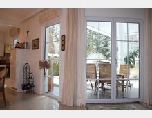 Wohnzimmer mit großen Fenstertüren, die auf eine Terrasse mit Holzmöbeln führen. Im Innenraum befinden sich helle Vorhänge, eine Vase mit Blumen und ein Weinregal; draußen ist eine schneebedeckte Gartenlandschaft sichtbar.