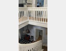 Treppenhaus in einem Wohnhaus mit weißen Holzgeländern und einer geschwungenen Treppe. Im unteren Bereich des Treppenhauses sind ein kleiner Holztisch, ein Spiegel und eine Lampe auf einem schwarz-weiß karierten Boden zu sehen; offene Türen führen in angrenzende Räume.