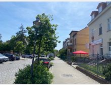 Eine gepflasterte Straße in einem Wohnviertel mit parkenden Autos, Reihenhäusern und Bäumen. Rechts im Bild befinden sich Eingänge zu Gebäuden mit Geländern, sowie ein roter Sonnenschirm vor einem Friseursalon.