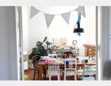 A cozy, cluttered workspace with a wooden table surrounded by mismatched chairs, shelves filled with books and art supplies, and green plants adding a touch of nature. A white wall features artwork, while a string of decorative pennants hangs from above, and a black pendant light shines over the table.