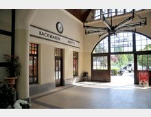 Innenbereich eines kleinen Bahnhofs mit hohen Fenstern und einem Rundbogenportal, das nach draußen führt. Links befinden sich eine Bäckerei und ein Café mit angeschriebenem Schild, daneben Blumen in einem Topf; rechts steht ein Werbeschild für Eis.