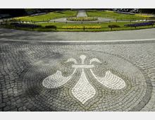 Steinpflasterplatz mit einem weißen Mosaikmuster in Form einer stilisierten Lilie im Vordergrund, dahinter ein runder Gartenbereich mit Blumenbeeten, Rasenflächen und umlaufenden Wegen. Im Hintergrund Bänke und weitere Blumenbeete.