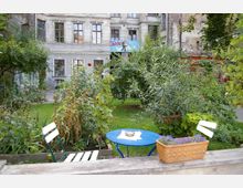Ein kleiner urbaner Garten mit einer Sitzgruppe aus blauen und weißen Gartenmöbeln, umgeben von dichtem Grün und Blumenbeeten. Im Hintergrund steht ein historisches Gebäude mit mehreren Fenstern und einer verwitterten Fassade.