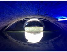 Gewölbter Tunnel mit Backsteinmauerwerk, beleuchtet von blauen Lampen. Im Vordergrund ist eine Wasserfläche zu sehen, die das Licht reflektiert, und im Hintergrund ein weiterer beleuchteter Durchgang.