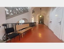 Flur in einem historischen Gebäude mit gewölbten Decken, weißen Wänden und einem rotbraunen Boden. An der linken Seite befinden sich Sitzgelegenheiten aus Holz und Metall, daneben ein kunstvolles Geländer, das eine Treppenöffnung flankiert.