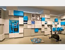 Ein moderner Ausstellungsraum mit interaktiven Elementen, darunter ein Heimtrainer, eine Gaming-Tanzmatte und eine Wand mit Exponaten wie Retro-Videospiel-Controllern und Infotafeln in blauen und weißen Kästen. Im Hintergrund befinden sich Bildschirme, Texte und historische Fotos, die Themen der Technologie und Interaktivität präsentieren.