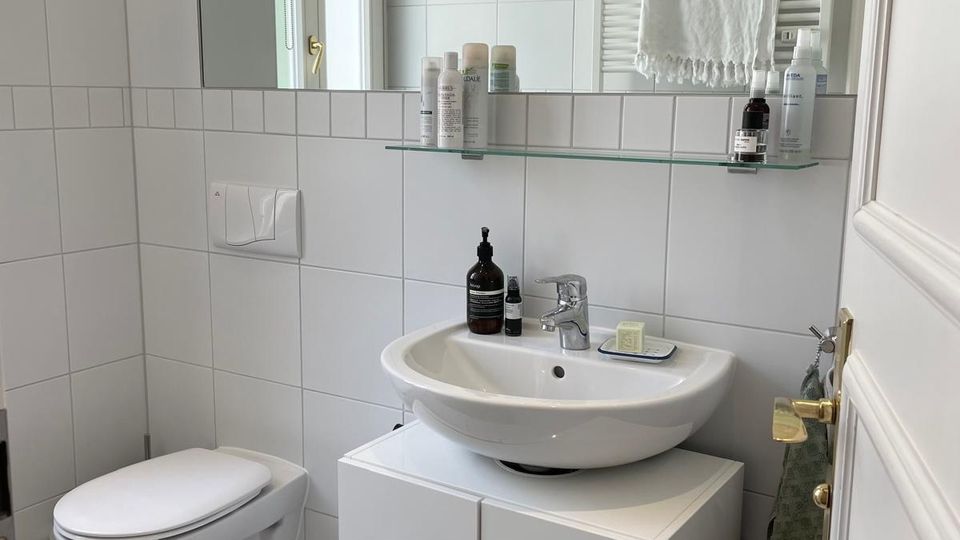 Helles, weiß gefliestes Badezimmer mit wandhängendem WC und Waschtisch auf einem Unterschrank. Über dem Waschbecken befindet sich ein breiter Spiegel mit Glasablage; im Hintergrund sind ein Handtuchheizkörper und ein Fenster zu sehen.