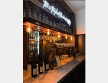 Innenansicht einer modernen Bar mit langer Theke aus Stein, hängenden Weingläsern und warmen Edison-Glühbirnen. Hinter der Theke stehen ein beleuchteter Getränkekühlschrank, Zapfhähne und ein Regal mit Neon-Schriftzug sowie gestapelten Gläsern.