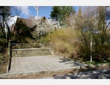 Ein gepflasterter Platz mit einer breiten Treppe, die zu einem Haus mit Satteldach und umgebender Vegetation aus Sträuchern und Bäumen führt; im Vordergrund stehen Straßenschilder mit den Beschriftungen „Arnold-Knoblauch-Ring“ und „Stölpchenweg“.
