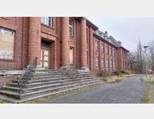 Backsteingebäude mit großen Säulen am Eingangsbereich und einer breiten Steintreppe mit kunstvollen Geländern, daneben ein Gehweg und kahle Bäume im Hintergrund. Die Fenster des Gebäudes sind teils mit Holzplatten verschlossen, was auf Verfall hindeutet.