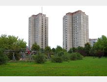 Zwei hohe Wohnhochhäuser aus Beton stehen nebeneinander, umgeben von grünen Bäumen und einer Grasfläche im Vordergrund, die einen Spielplatz mit Holzelementen enthält. Der Himmel ist bedeckt.