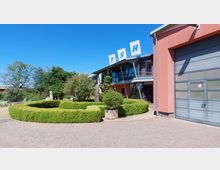 Ein moderner Gebäudekomplex mit einer auffälligen rosa Fassade, einem hohen Metalltor und großen Glasflächen. Im Vordergrund befindet sich eine gepflegte Gartenanlage mit grünen Hecken und kleinen Bäumen, umgeben von einem gepflasterten Weg; darüber hinaus befinden sich oben auf dem Gebäude die Buchstaben "TSH".