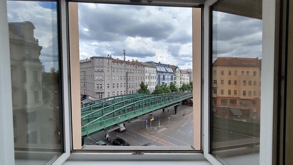 Blick aus einem geöffneten Fenster auf eine städtische Straße mit einer grünen Hochbahnbrücke und mehreren mehrstöckigen Wohn- und Geschäftshäusern. Im Hintergrund ist ein bewölkter Himmel über den Gebäudefassaden zu sehen.
