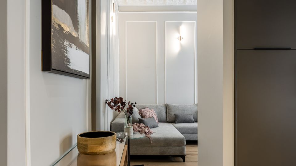 Blick durch einen schmalen Flur mit hellem Holzboden und weißer Wandvertäfelung in ein Wohnzimmer mit grauem Ecksofa. An der linken Wand hängt ein großes abstraktes Bild über einer schwebenden Glas-Konsole, rechts steht ein hoher dunkler Schrank; an der Decke sind Stuckleisten und eine Wandleuchte zu sehen.