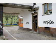 Ansicht eines Straßenecks mit einem Gebäude im Stil der Nachkriegsarchitektur. Im Vordergrund ist ein Eingang mit graffitibeschrifteten Wänden zu sehen, daneben ein Schaufenster mit orangefarbener Werbung. Im Hintergrund ein Wohnhaus in grüner Fassade sowie parkende Autos.