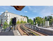 Eine erhöhte U-Bahn-Station mit gebogenen Gleisen, flankiert von historischen Wohngebäuden im Gründerzeitstil und umgeben von grünen Bäumen. Im Hintergrund sind klare Himmel und weitere städtische Elemente zu sehen.