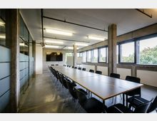 Moderner Konferenzraum mit einer langen rechteckigen Tischanordnung, umgeben von schwarzen Stühlen. Der Raum hat große Fenster, die Tageslicht hereinlassen, Sichtbetonwände, eine Glaswand mit mattem Design und eine Wand mit einem großen Bildschirm.