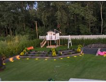 Garten mit angelegtem Rasen, Büschen und Steinterrassen, im Hintergrund ein Spielhaus auf Stelzen mit einer Rutsche und Schaukeln. Links und rechts des Gartens ist dichter Wald zu sehen, im Vordergrund befinden sich beleuchtete Beete und ein Poolbereich.