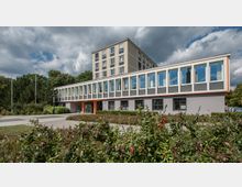 Ein modernes Verwaltungsgebäude mit grauer Fassade, großen rechteckigen Fenstern und einem orangefarbenen Eingangsbereich. Im Vordergrund befinden sich grüne Büsche und ein gepflasterter Weg, im Hintergrund sind Bäume und ein bewölkter Himmel zu sehen.