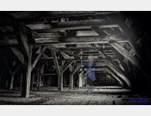 Ein dunkler Dachboden mit einer alten Holzbalkenkonstruktion, Staubpartikeln in der Luft und einem schwach erkennbaren, halbtransparenten menschlichen Umriss in der Mitte der Szene.