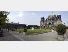 Weitläufiger Platz mit gepflasterten Wegen und Grünflächen, im Hintergrund der Berliner Dom mit seinen markanten Kuppeln und das Alte Museum mit Säulenfassade. Auf dem Rasen sind Menschen zu sehen, eine Fontäne sprüht Wasser in der Mitte.