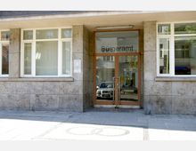 Eingang eines Bürgeramts mit einer Glastür in einem Gebäude mit hellem, steinverkleidetem Fassadenmaterial. Links und rechts der Tür befinden sich große Fenster, die den Innenraum teilweise sichtbar machen.