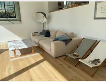 Helles, modern eingerichtetes Wohnzimmer mit hellem Holzboden, beigem Ecksofa und niedrigem Couchtisch aus Holz. An der Wand hängen gerahmte Bilder, daneben stehen zwei klappbare Liegestühle; eine Stehlampe und eine halbhohe Trennwand mit Ablage strukturieren den Raum.