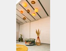 Moderner Innenraum mit einer cremefarbenen Holzwand im Hintergrund, einer gefliesten Decke mit warm leuchtenden Glaskugellampen sowie dekorativen Elementen wie einer großen Vase mit Pampasgras, einer farbigen geflochtenen Sitzgelegenheit und einem gelben Teppich.