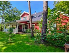 Rot gestrichenes Haus mit weißem Holzverandadach, großen Fenstern und Satteldach, umgeben von einer gepflegten Rasenfläche, Bäumen und dichtem Grün. Der Himmel ist klar und blau, was auf eine sonnige Umgebung hindeutet.