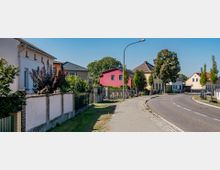 Wohnstraße in einer ländlichen Umgebung mit Einfamilienhäusern, teils mit bunten Fassaden. Die Straße ist leicht geschwungen, mit gepflastertem Gehweg und vereinzelten Straßenlaternen, umgeben von gepflegten Gärten und jungen Bäumen.