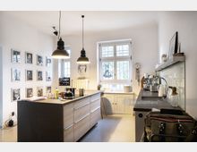 Helle Wohnküche mit modernem Design, zentraler Kücheninsel aus Holz und schwarzen Arbeitsplatten. An der Wand hängen Porträtfotos, und große Fenster lassen viel Tageslicht herein; im Hintergrund befinden sich eine Arbeitsfläche mit Spüle sowie eine Kaffeemaschine.