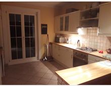 Eine helle Küche mit gelben Wänden, einer Arbeitsplatte aus Holz und einem Fliesenboden. Die Küche verfügt über Hängeschränke mit Glaseinsätzen, einen Gasherd mit Dunstabzugshaube, sowie eine gläserne Doppeltür, die nach außen führt.