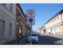 Straßenansicht mit Wohn- und Geschäftshäusern auf beiden Seiten, teilweise verputzt oder mit Backsteindetails. Im Vordergrund stehen Verkehrsschilder für eine Tempo-30-Zone und Halteverbot, daneben ein parkendes Auto und eine Laterne.