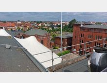 Blick auf ein historisches Backsteingebäudeensemble mit mehreren Gebäuden, darunter ein rundes Bauwerk mit Kuppeldach. Im Vordergrund sind weiße, zeltartige Überdachungen und ein Gebäudedach mit Geländer sichtbar; im Hintergrund erstreckt sich eine Kleinstadt mit roten Dächern und vereinzelten Grünflächen.