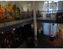 Innenraum eines Museums mit Ausstellungsstücken, darunter Marionetten und farbenfrohe Figuren, die auf einem erhöhten Laufsteg präsentiert werden. Das zweistöckige Gebäude hat hohe Fenster, einen weißen Pfeiler und Wandtafeln mit Informationen sowie eine beleuchtete Ausstellungsfläche im unteren Bereich.