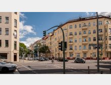 Straßenecke in einer Stadt mit mehrstöckigen Wohngebäuden im Altbaustil, farblich in Beige und Rot gehalten, sowie mehreren Balkonen. Im Vordergrund befindet sich eine Kreuzung mit Verkehrsschildern, Ampeln, Autos und einem Fahrradfahrer, umgeben von Bäumen und Gehwegen.