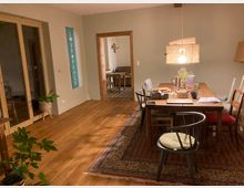 Esszimmer mit Holzboden und großem Holztisch, umgeben von verschiedenen Stühlen. Im Raum stehen grüne Pflanzen, eine Pendelleuchte aus Stoff hängt über dem Tisch, und im Hintergrund ist eine offene Tür zu einem weiteren Raum sichtbar.