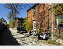 Straßenansicht mit Kopfsteinpflaster, flankiert von roten Backsteingebäuden. Vor einem der Gebäude stehen ein Kleinwagen, ein Motorrad und ein Motorrad mit Beiwagen; Bäume werfen Schatten auf die Gebäude und die Straße.