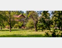 Backsteinhaus mit rotem Ziegeldach, umgeben von einer weitläufigen Wiese und mehreren Bäumen, in einer ländlichen, bewaldeten Umgebung. Die Fassade des Hauses ist teilweise von Vegetation verdeckt.