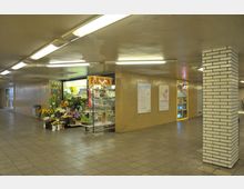 Eine unterirdische Passage mit gelblich beleuchteten Wänden, einem Blumenladen auf der linken Seite und einer gefliesten Säule im Vordergrund. Der Boden besteht aus grauen Fliesen, und an den Wänden hängen Aushänge und Schaufenster.