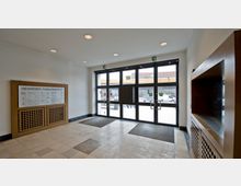 Foyer eines Gebäudes mit großen Glas-Eingangstüren, die nach außen auf eine Straße mit geparkten Fahrzeugen führen. Links an der Wand ein „Wegweiser Familiengericht“, rechts eine hölzerne Vitrine, der Boden ist mit hellen Fliesen gestaltet.