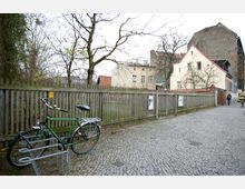 Gepflasterter Gehweg entlang eines Holzzauns, dahinter wenige laublose Bäume und ein weißes Gebäude mit Satteldach. Im Vordergrund steht ein grünes Fahrrad in einem Fahrradständer, rechts im Bild spazieren zwei Personen.