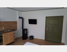 Ein schlicht eingerichteter Raum mit weiß gestrichenen Wänden, einem Kaminofen mit schwarzem Rohr, einem grünen Türrahmen mit passender Tür, einem an der Wand montierten Flachbildfernseher und einer kleinen Küchenzeile mit Holzfronten und heller Arbeitsplatte. Der Boden besteht aus Holzoptik-Laminat.