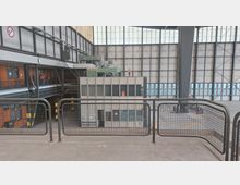 Ein großzügiger, industrieller Innenraum mit einem hohen Dach aus Lichtpaneelen, großen Metallstrukturen und einer kleinen Glas- und Metallkabine im Zentrum. Im Hintergrund befinden sich Gittertore und Fensterreihen, Personen in orangenen Warnwesten sind erkennbar.