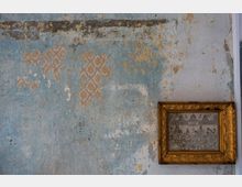 Abblätternde, blaue Wandfarbe mit sichtbaren goldenen und weißen Musterspuren auf einer alten Wand; rechts hängt ein kleines gerahmtes Bild mit einer kunstvollen, goldfarbenen Rahmenverzierung.