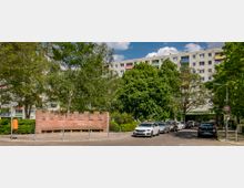 Straßenszene mit einem Denkmal aus rotem Sandstein im Vordergrund, das Reliefs zeigt, umgeben von Bäumen und Sträuchern. Im Hintergrund sind Wohnblöcke mit bunten Fensterläden und ein Straßenschild mit dem Schriftzug "Charlottenboulevard" zu sehen; entlang der Straße parken mehrere Fahrzeuge.