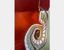 Spiraltreppe mit geschwungenem Geländer aus Metall, die sich in einem mehrstöckigen Treppenhaus nach unten windet. Die Wände sind in Rot gehalten, während die Stufen helle Farben aufweisen.