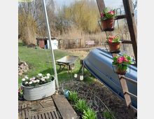Gartenbereich mit einem umgedrehten blauen Boot, das auf einem Gestell liegt, daneben eine Holzleiter mit Blumenkübeln. Im Hintergrund ein Steg mit Bank an einem Schilfgesäumten Gewässer, vorne eine Schubkarre und ein Pflanzgefäß mit Blumen.